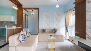 A modern living room with a sofa, a round gold-framed side table, and a kitchen area visible through an open doorway.