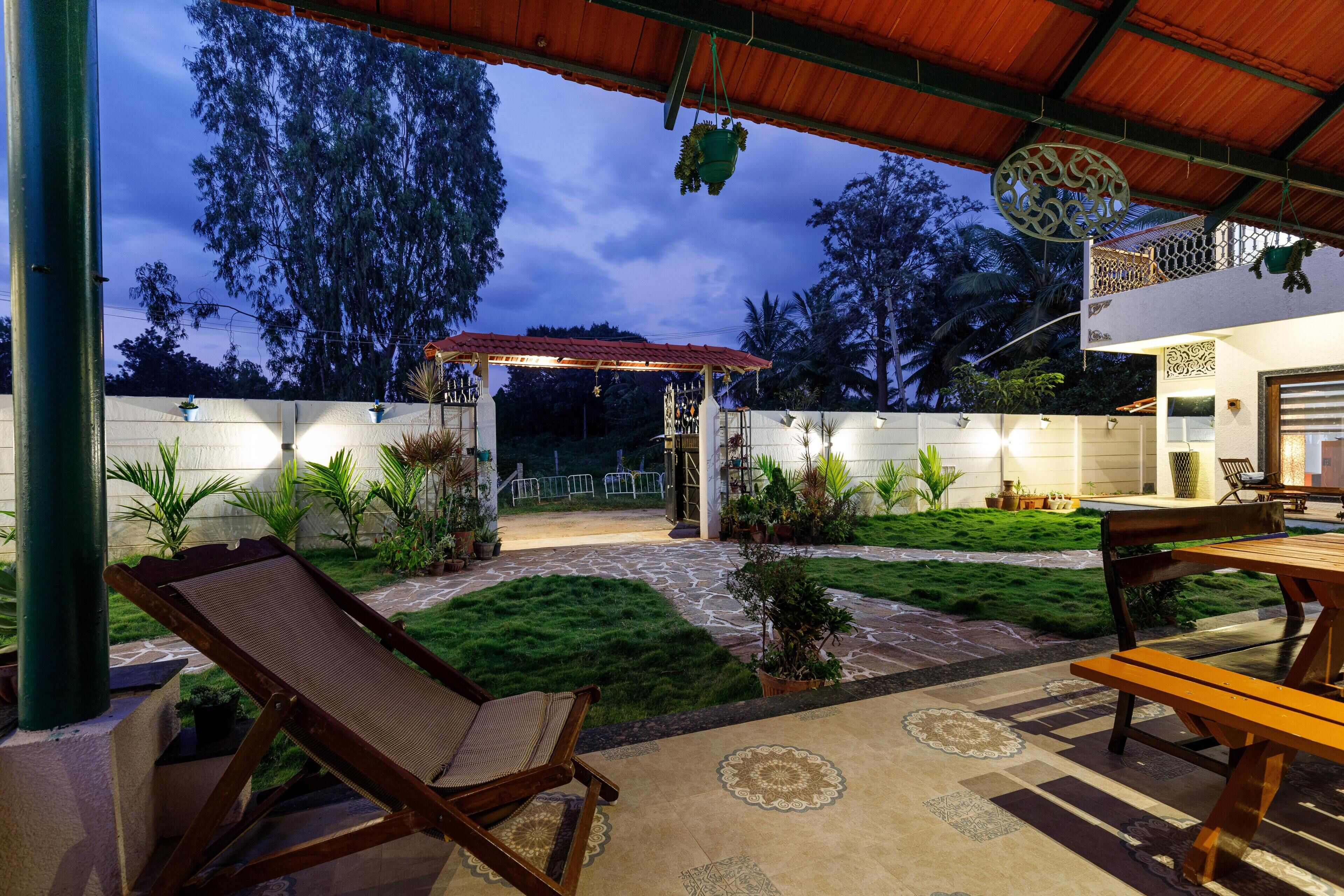 A covered outdoor patio with a lounge chair, a wooden table, and a small garden area with potted plants and a stone pathway.