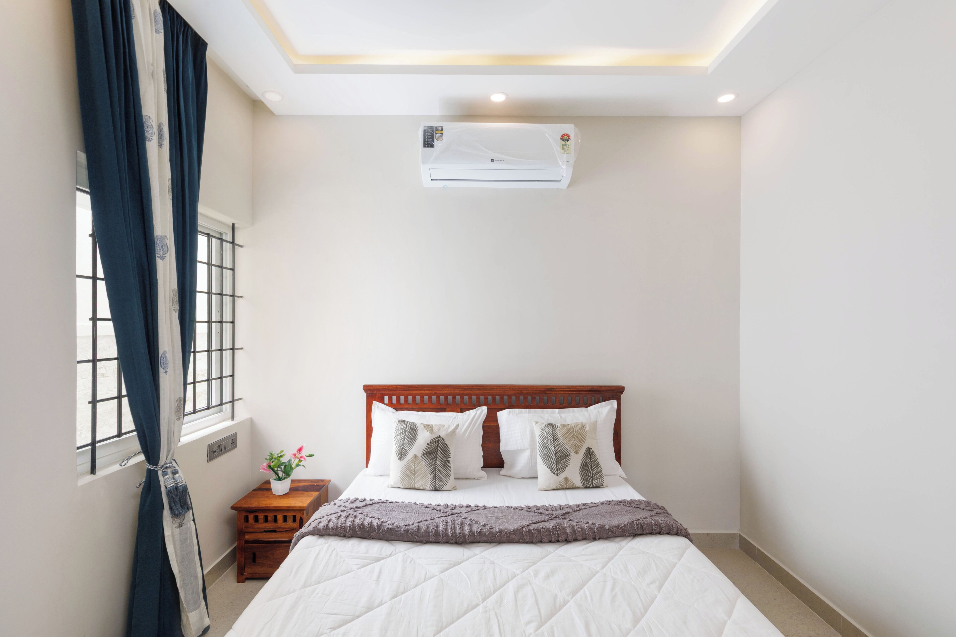 Un dormitorio con cama, mesita de noche, planta en maceta, ventana con cortinas y equipo de aire acondicionado.