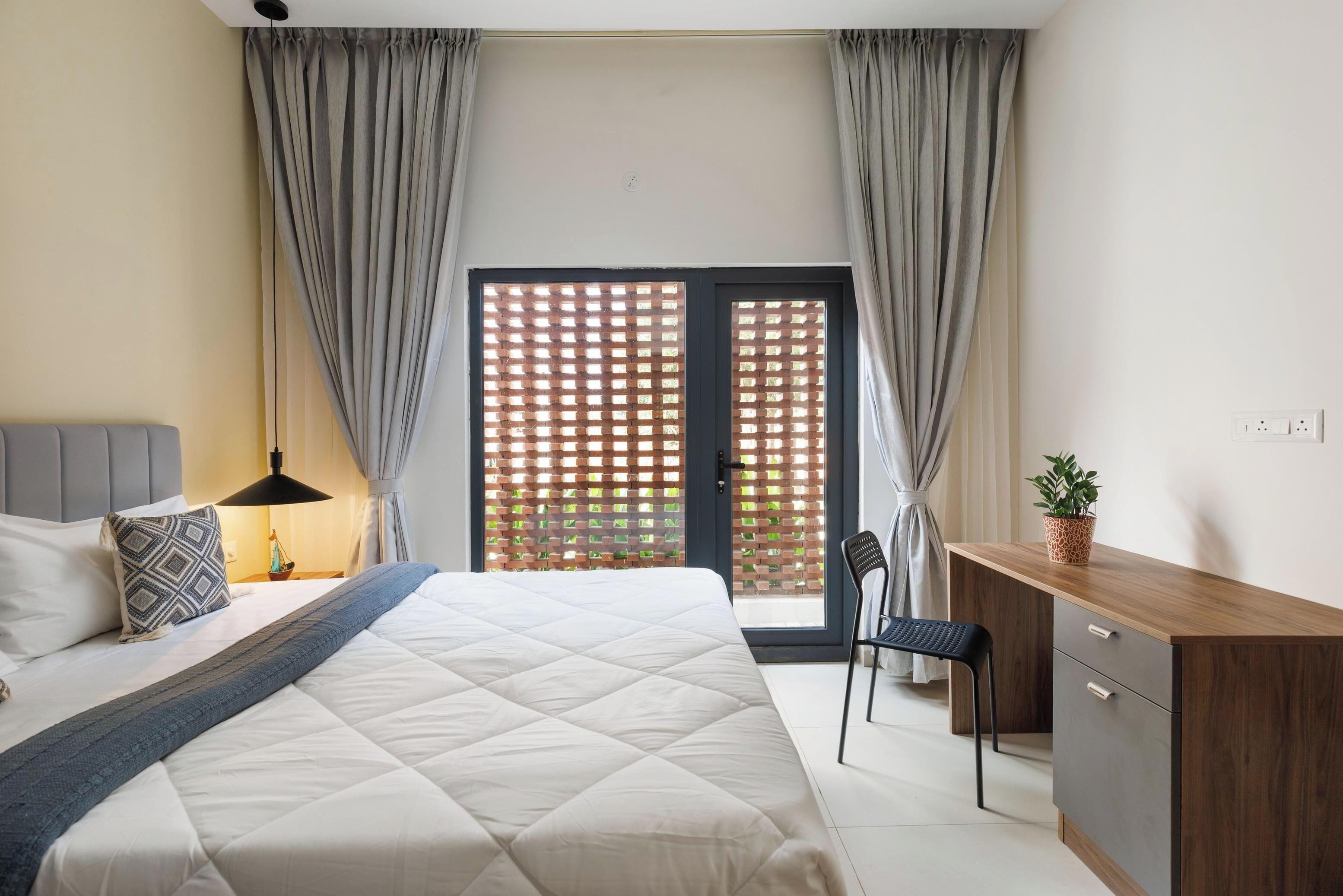 A bedroom with a bed, bedside table, lamp, desk, chair, and a view through a window with curtains.