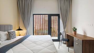 A bedroom with a bed, bedside table, lamp, desk, chair, and a view through a window with curtains.