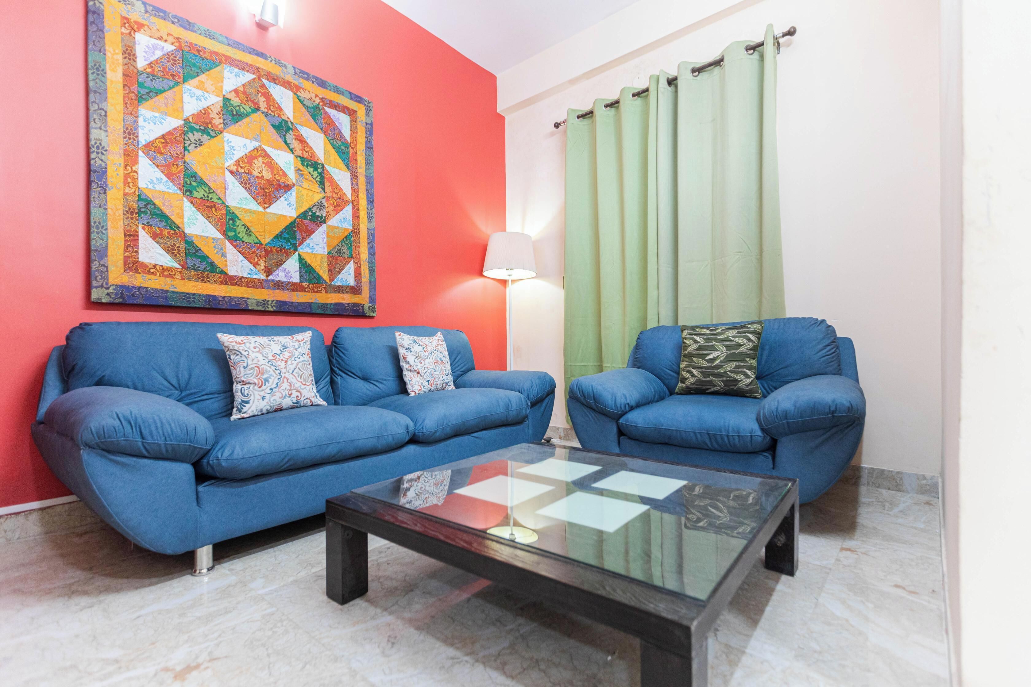 A living room with two blue sofas, a glass coffee table, a colorful quilt on the wall, and a green curtain.
