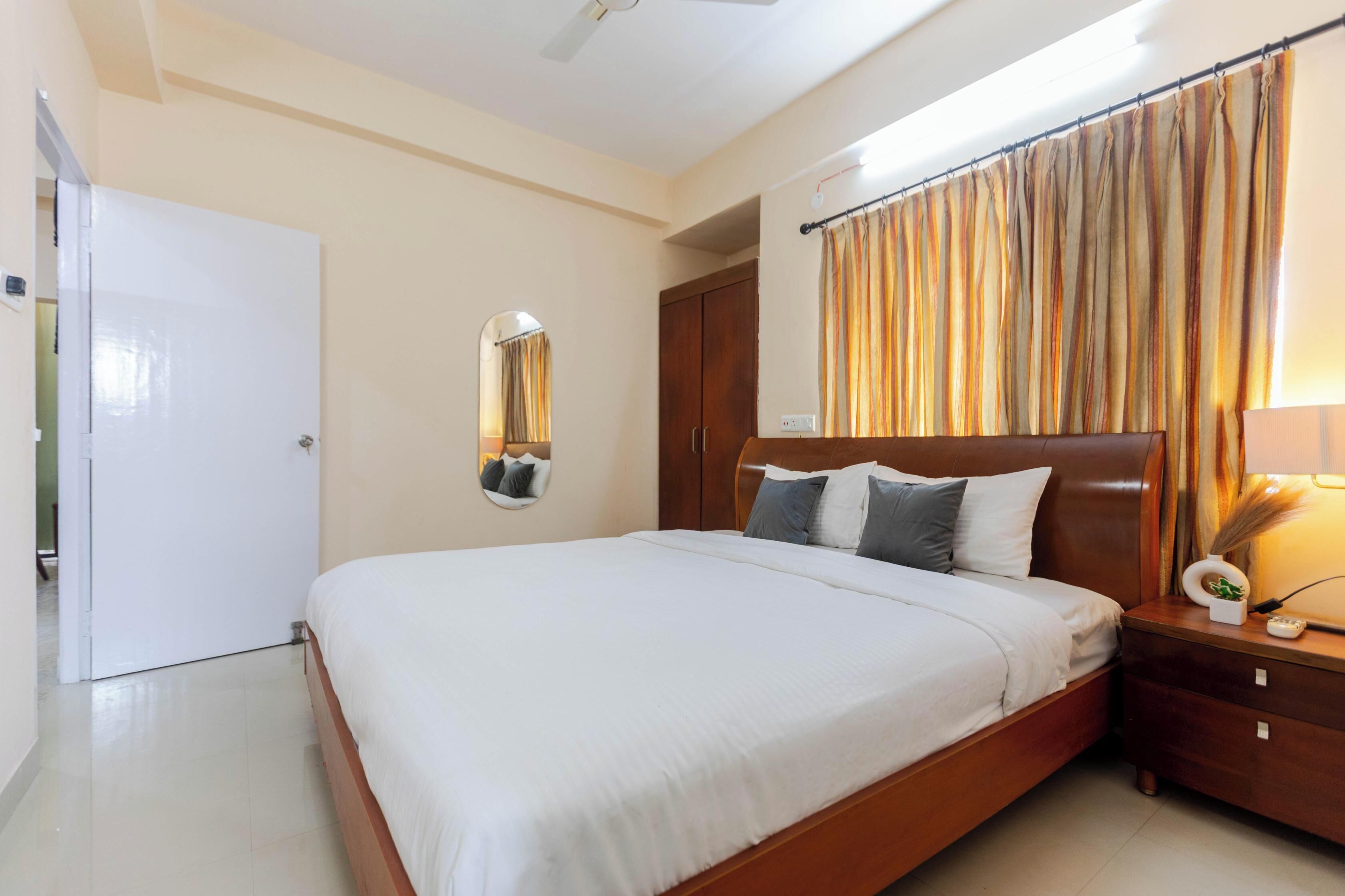 Um quarto com uma cama, mesa de cabeceira, espelho, guarda-roupa e cortina.