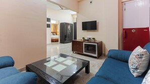 A living room with a glass coffee table, blue sofas, a TV mounted on the wall, a red door, and a kitchen area in the background. - Elara Residences by Jadecaps2bhknr IIM Kolkata (Alipur)