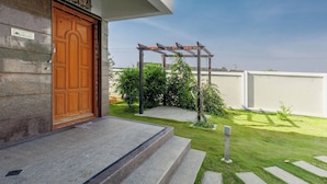 A building entrance with a wooden door, steps leading up to a porch, a metal pergola, and a well-maintained lawn. - Jhilmil Escape Pool Garden 1hr Koramangala HSR (Hosur)