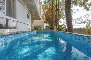 Piscina con mosaicos azules, rodeada de árboles y un edificio con grandes ventanales.