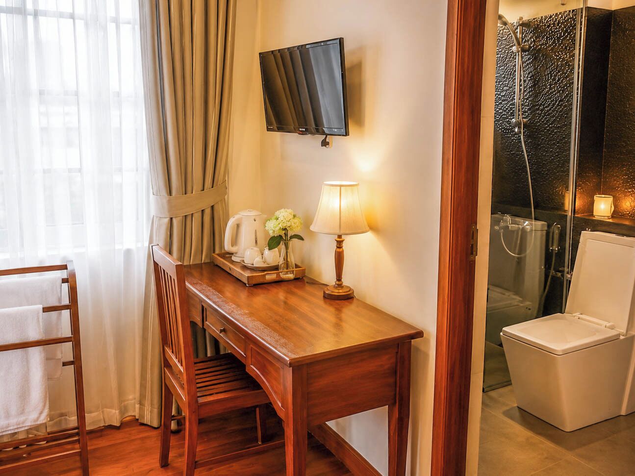 Une chambre d’hôtel avec un bureau en bois, un téléviseur fixé au mur, un espace douche et des toilettes blanches.