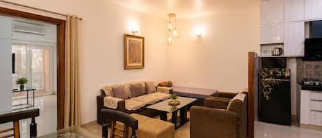 A modern living room with a glass dining table, sofa, and armchairs, a ceiling fan, and a kitchen with a refrigerator and microwave.