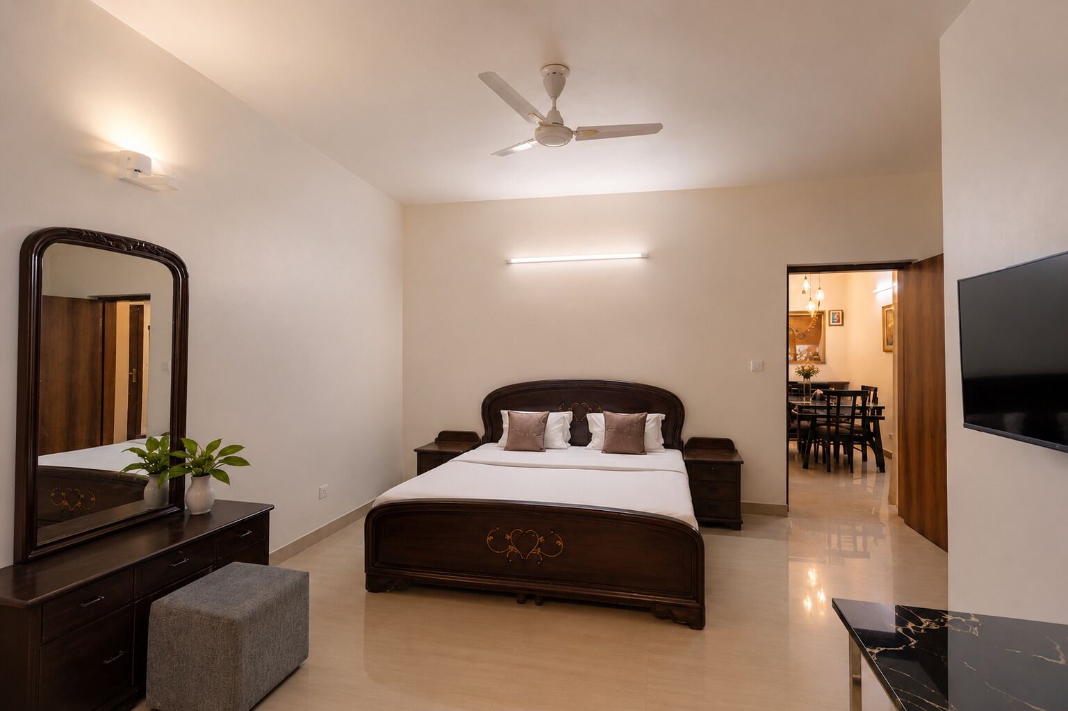 A bedroom with a bed, bedside tables, a mirror, a ceiling fan, a TV, and a dining area.