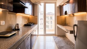 A modern kitchen with dark wood cabinets, stainless steel appliances, and a large window providing a view of buildings outside. - Aravalli by Red Olive4bhk10min Apollocybercity (Gurugram)