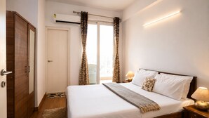 A bedroom with a bed, bedside tables, a wardrobe, a window with curtains, and an air conditioning unit. - Aravalli by Red Olive4bhk10min Apollocybercity (Gurugram)