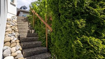 Eine Steintreppe führt zu einem Gebäude, umgeben von Grünpflanzen und einer Holzhandläufe.