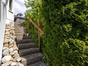 Un escalier en pierre menant à un bâtiment, entouré de verdure et doté d’une rambarde en bois.