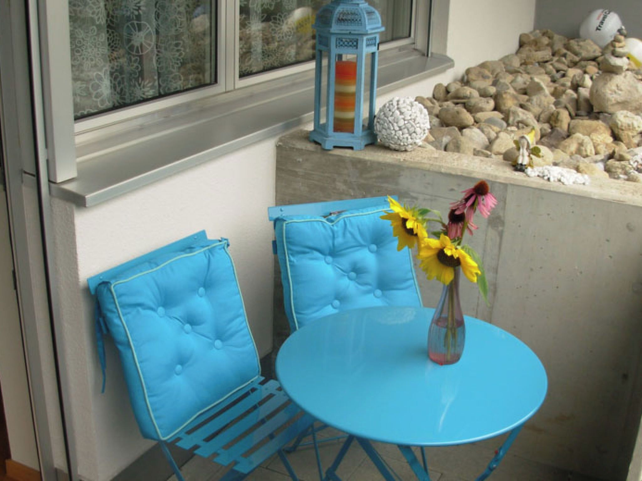 Un coin aménagé avec deux chaises bleues, une petite table ronde, un vase de fleurs, une lanterne et un jardin de rochers.