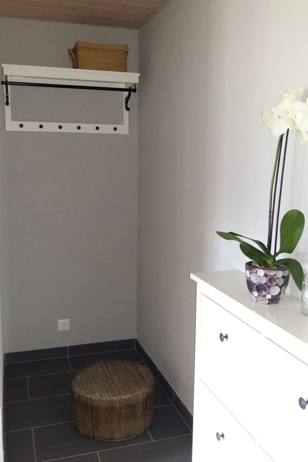 Ein Badezimmer mit einem weißen Schrank, einem Korbstuhl, einem Regal mit Korb und einer Topfpflanze mit weißen Blüten.