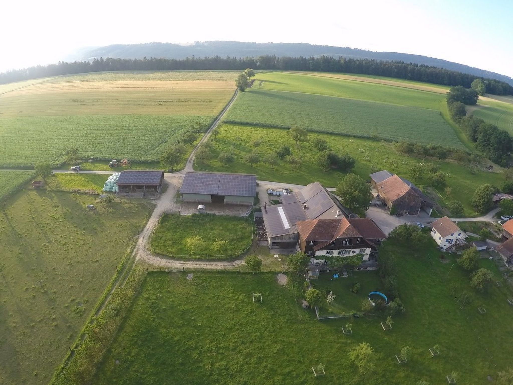 A rural property with multiple buildings, including a large house with a sloped roof, a barn, and several smaller structures. There is a fenced area with a solar panel array, and the property is surrounded by expansive fields.