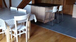 A dining area with a table and chairs, a kitchen with a bar, and a wooden ceiling. - Appartement - Serena, (Courtetelle), by Interhome (Courtételle)