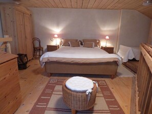 A bedroom with a large bed, two bedside lamps, a wicker chair, and a wooden dresser. - Studio - Le Râtelier, (Coeuve), by Interhome (Coeuve)