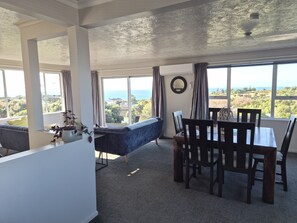 Ein Zimmer mit Esstisch, Stühlen, einer Couch und großen Fenstern mit Meerblick.