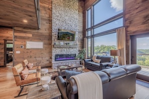 A spacious living room with a stone fireplace, leather sofas, and a large window offering a view of the outdoors. - 4 King Suites | Indoor Pool + Mtn Views (Sevierville)