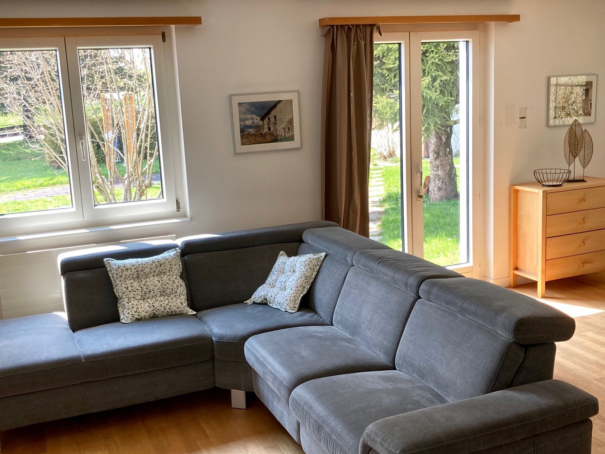 Ein modernes Wohnzimmer mit einem grauen Ecksofa, einer Holzkommode und einem großen Fenster mit Blick auf Grünflächen.