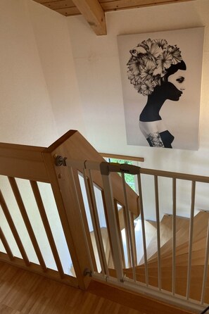Een houten trap met een leuning, leidend naar een kamer met een witte wand en een kunstwerk met een bloemistisch ontwerp.