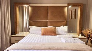 Habitación de hotel con una cama grande, un cabecero de madera, una mesita de noche con un reloj y un espejo.