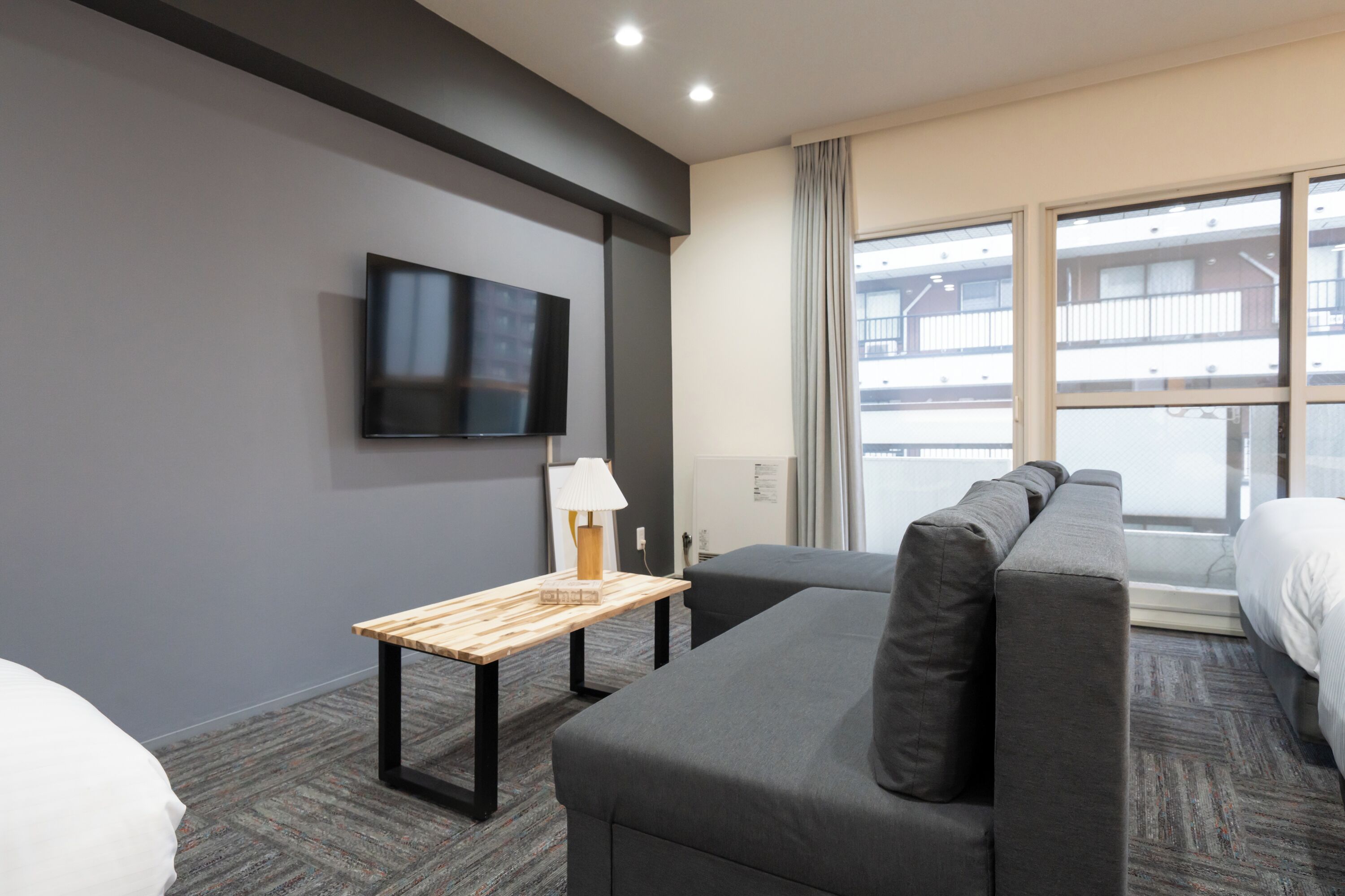 Habitación de hotel con sofá gris, una pequeña mesa de madera con lámpara, televisor de pantalla plana montado en la pared y un amplio ventanal con vista al exterior del edificio.