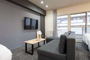 Habitación de hotel con sofá gris, una pequeña mesa de madera con lámpara, televisor de pantalla plana montado en la pared y un amplio ventanal con vista al exterior del edificio.