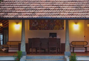 A covered outdoor seating area with a tiled roof, two wooden chairs, a bench, and a wall-mounted computer monitor.