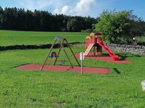 Ein Spielplatz mit Schaukel und Rutsche, umgeben von einer grünen Wiese und Bäumen.