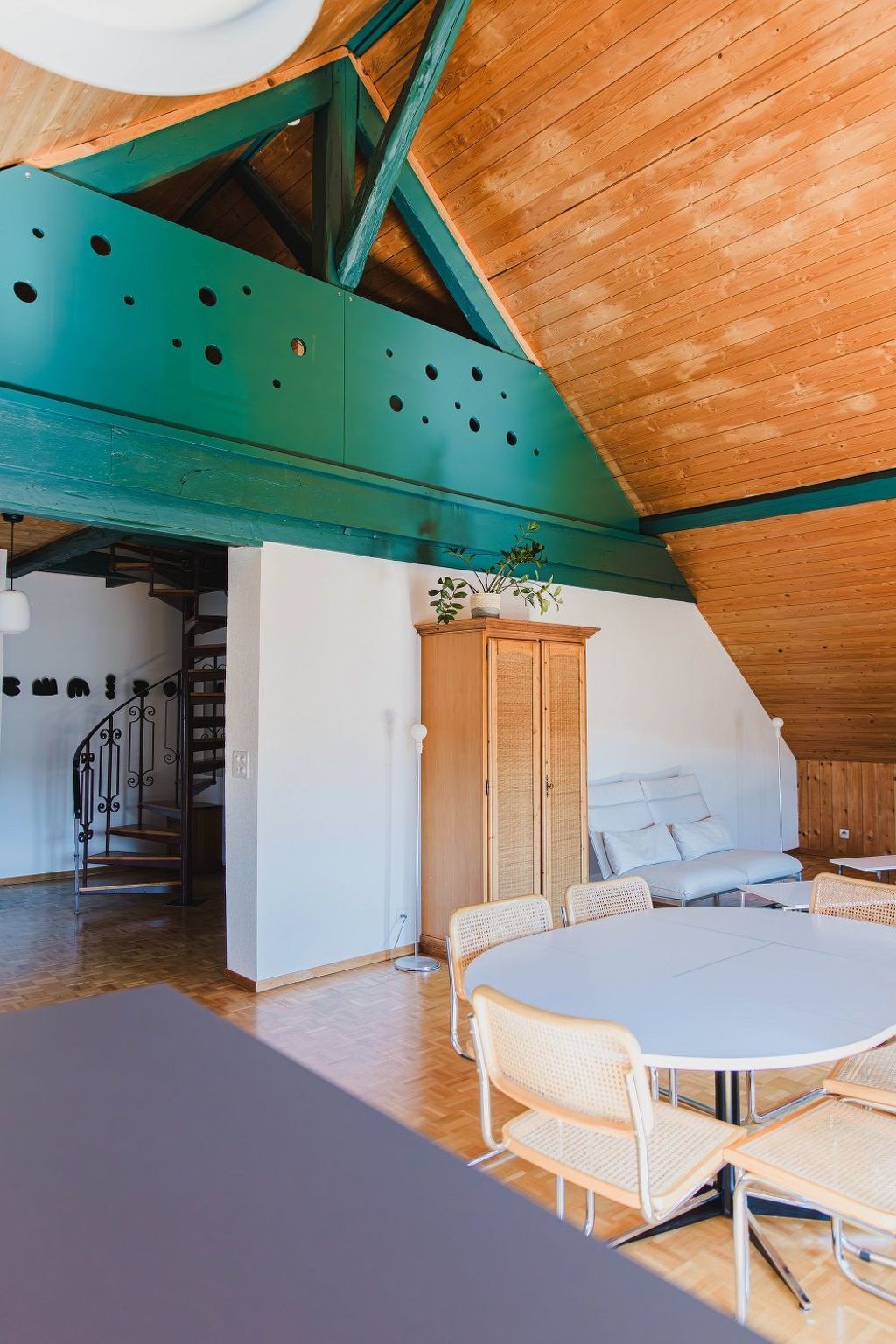 Ein Zimmer mit hoher Decke, Holzbalken, einer Treppe, einem Essbereich mit rundem Tisch und Stühlen sowie einem Bett mit Kissen.