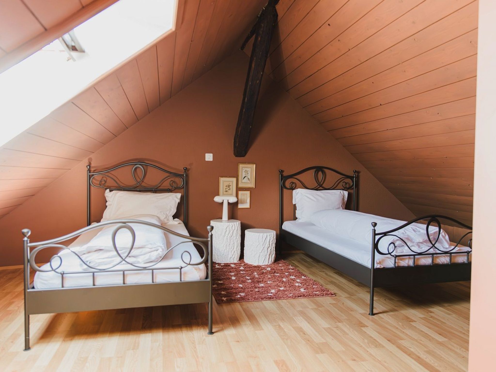 Ein quadratisches Schlafzimmer im Dachgeschoss mit zwei Metallbetten, einer Holzbalkendecke und einem roten Teppich.
