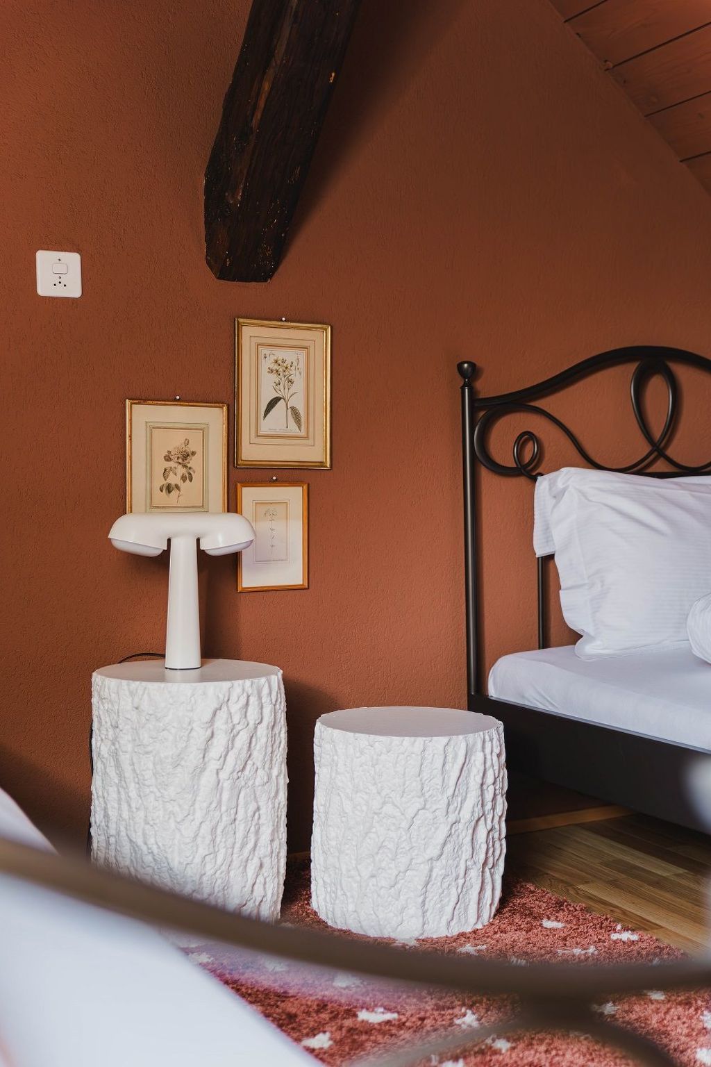 A bedroom with a bed, two white textured stools, and framed botanical prints on the wall.