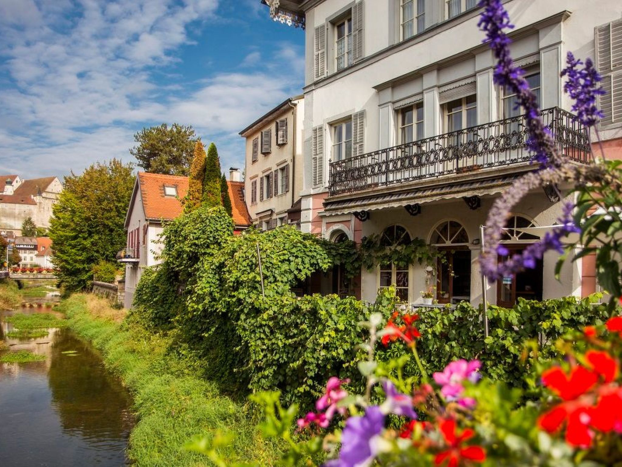 Ein Gebäude am Flussufer mit einem Balkon, umgeben von üppigem Grün und Blumen.
