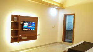 A hotel room with a mounted TV displaying various streaming services, a wooden shelf unit, a bed with a brown blanket, and a bathroom visible through a glass door. - Vrindavan Guest house  (Ambegaon)