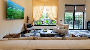 A modern living room with a large flat-screen TV, a sofa, a coffee table, and a view of greenery through the window. - Orfeas Villas 2 Canggu Villa 2BR Pool (Canggu)