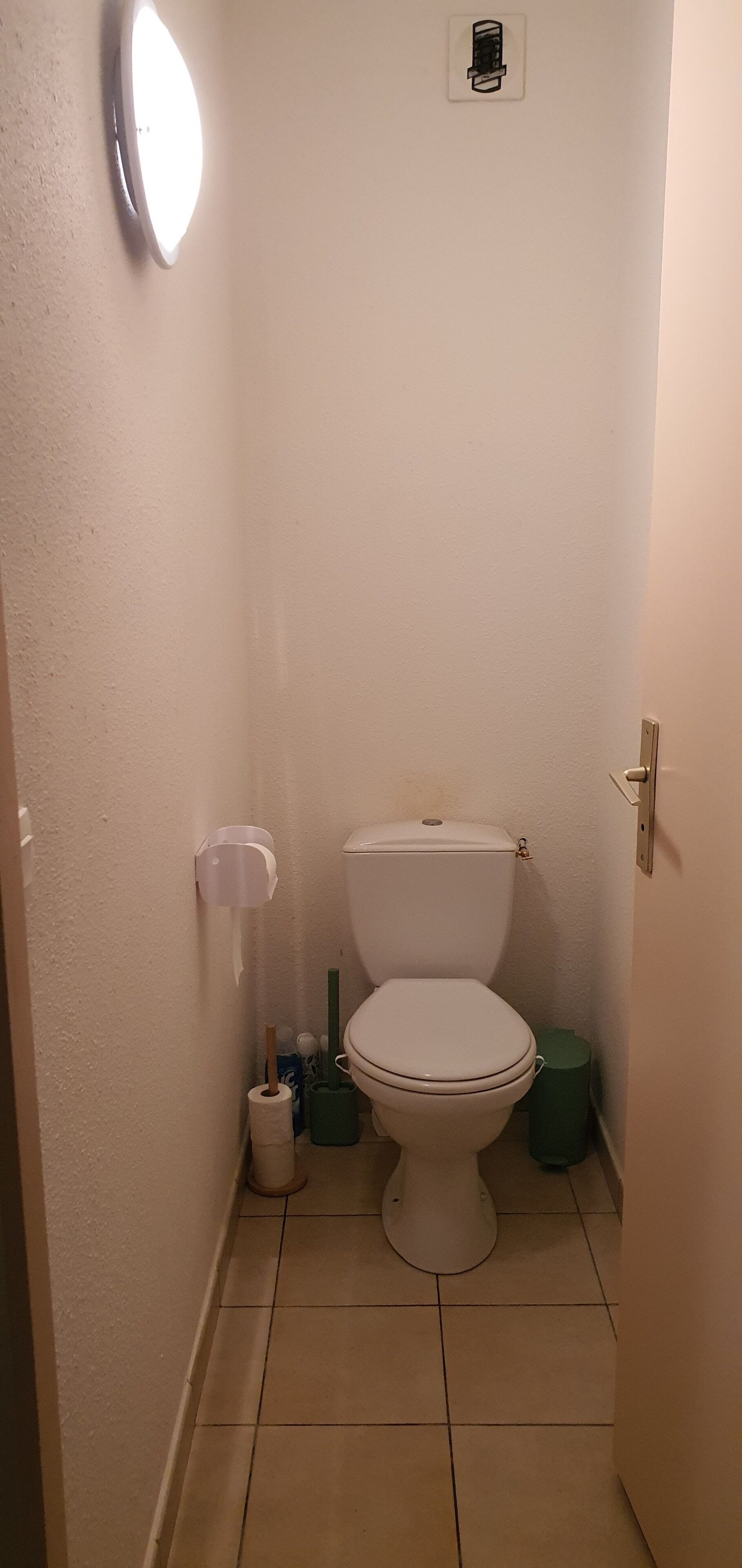 Ett litet, smalt badrum med en toalett, en vägghängd toalettpappershållare, en papperskorg och en grön rengöringshink.