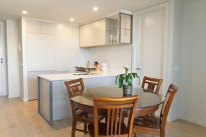 A modern kitchen with white cabinets, a central island, and a dining table with chairs. - Spacious New 2 BDR with City Views (Northcote)