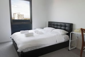 A single bed with white linens and a black headboard in a room with a window and a small desk. - Spacious New 2 BDR with City Views (Northcote)