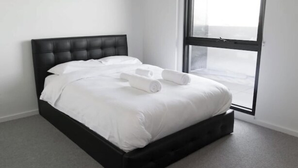 Une chambre moderne avec une tête de lit noire, du linge de lit blanc et une vue dégagée sur l’extérieur grâce à une grande fenêtre.