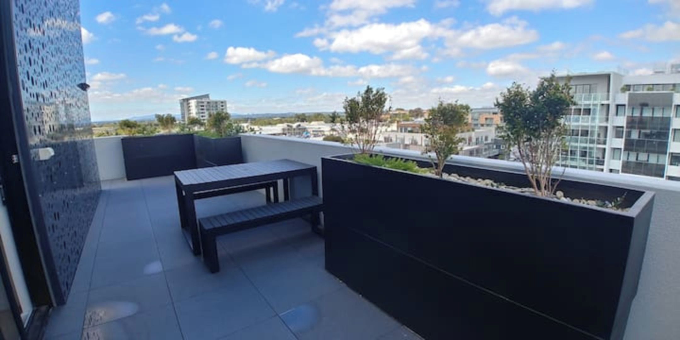 A rooftop terrace with a table and benches, surrounded by a low wall with plants.