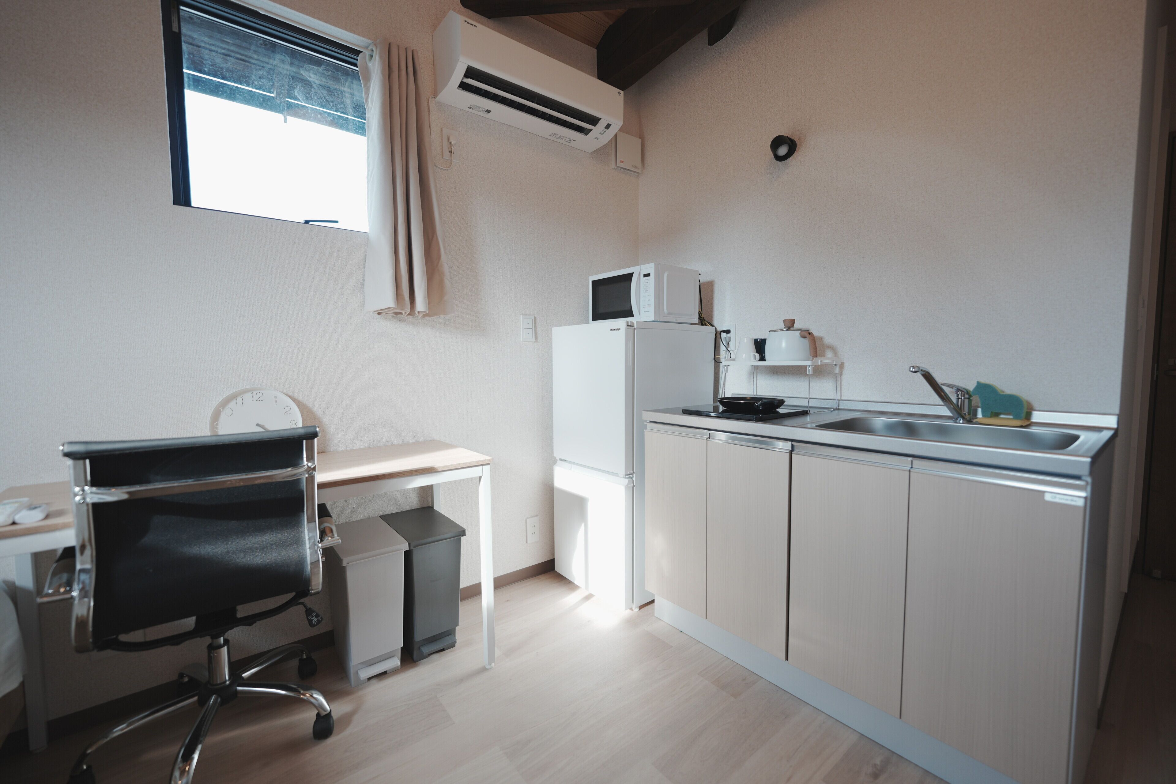 A compact kitchen with a microwave, sink, and a small refrigerator. There's a chair and a desk with a clock.