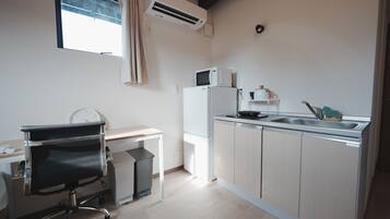A compact kitchen with a microwave, sink, and a small refrigerator. There's a chair and a desk with a clock.