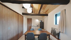 A room with a wooden dining table, four chairs, a kitchen area with a microwave and refrigerator, and a window with curtains.