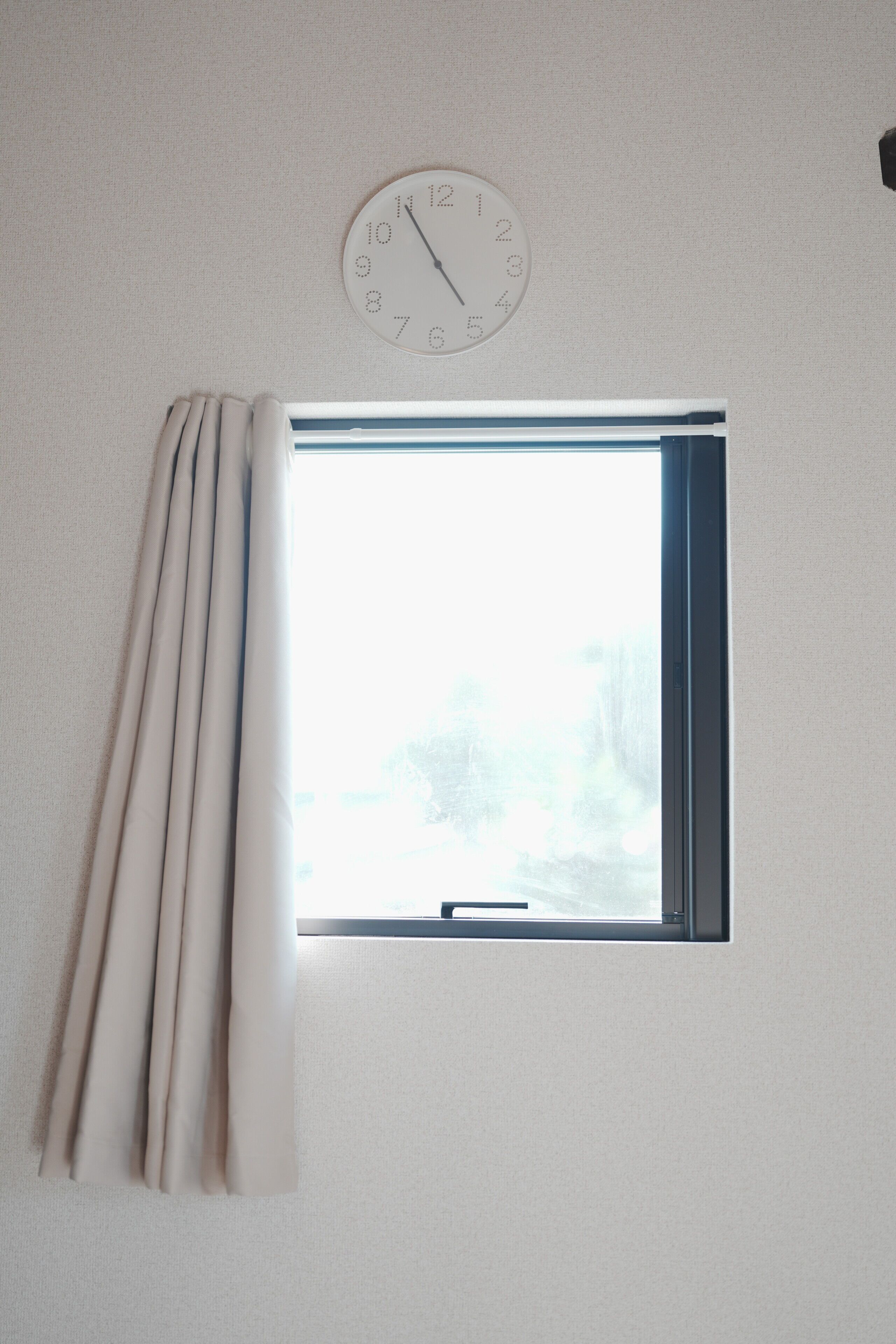 A window with curtains, a wall clock, and a view of trees.