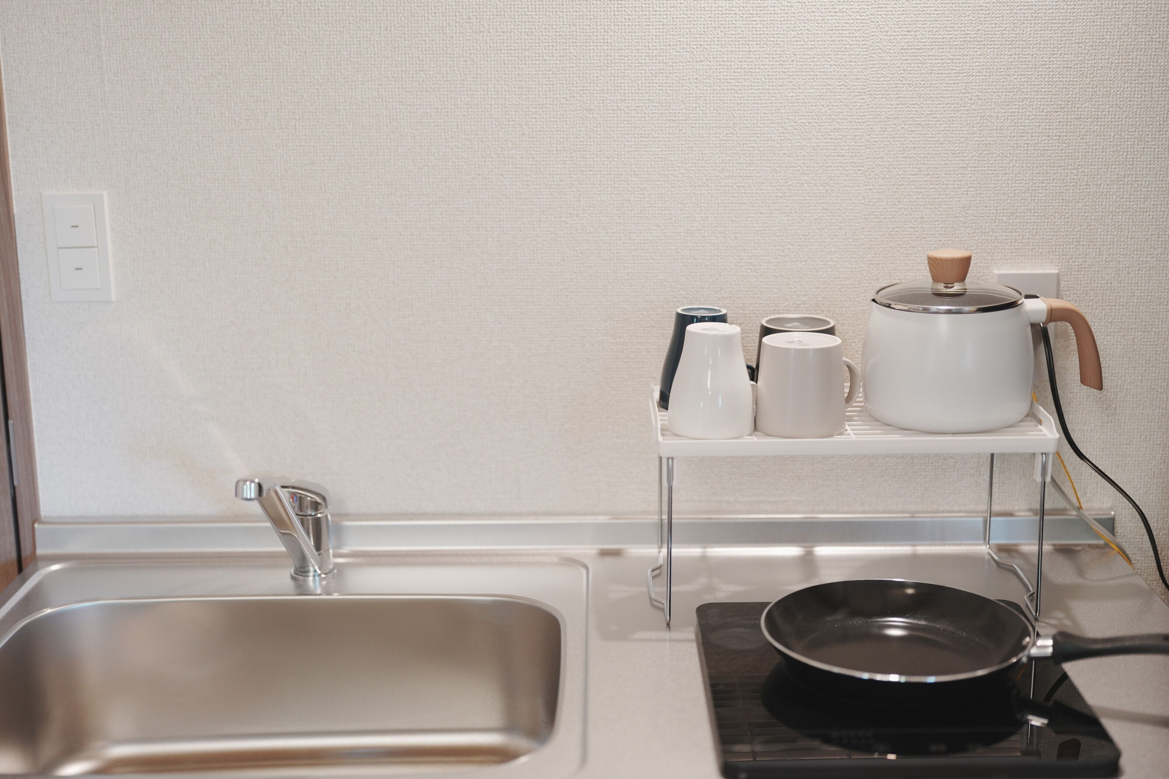 A kitchen with a stainless steel sink, a kettle, a coffee maker, and a frying pan on a portable stove.