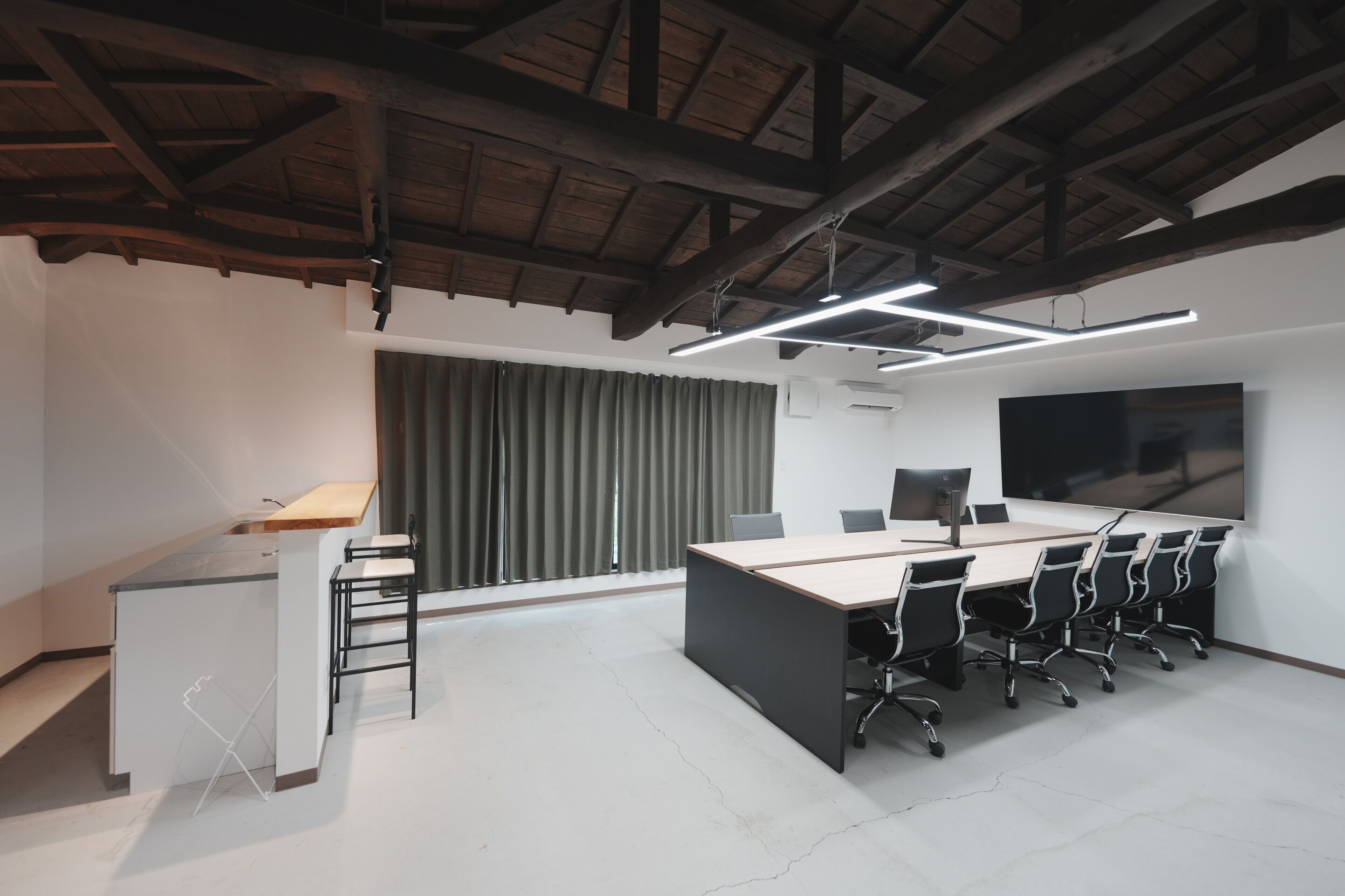 A modern office space with a large desk, multiple chairs, a large screen, and a window with curtains.