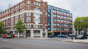 Un edificio de varios pisos con fachadas de ladrillo y vidrio, una calle al frente y coches estacionados.