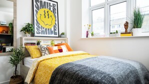 Un dormitorio con cama, ventana, estantería con libros y una obra de arte en la pared.